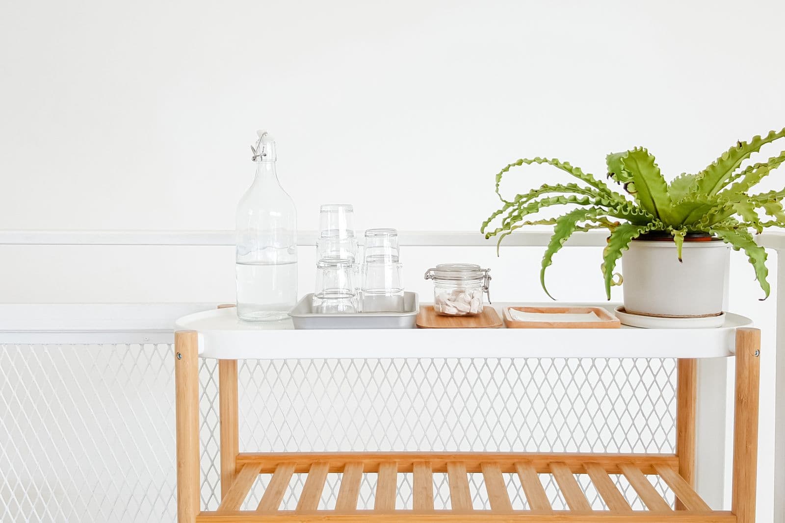 A bright two-tier cart holding a tray of glasses, a bottle and a plant