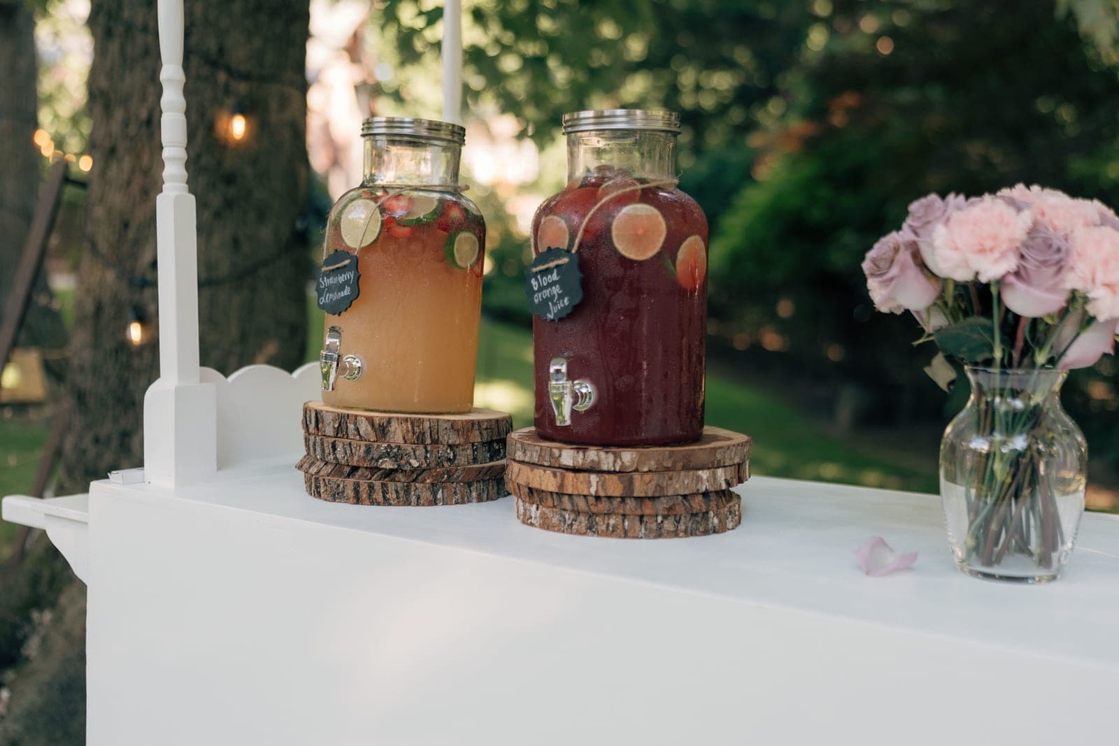 Two glass drinks dispensers and pink roses on a white serving cart in a sunlit garden