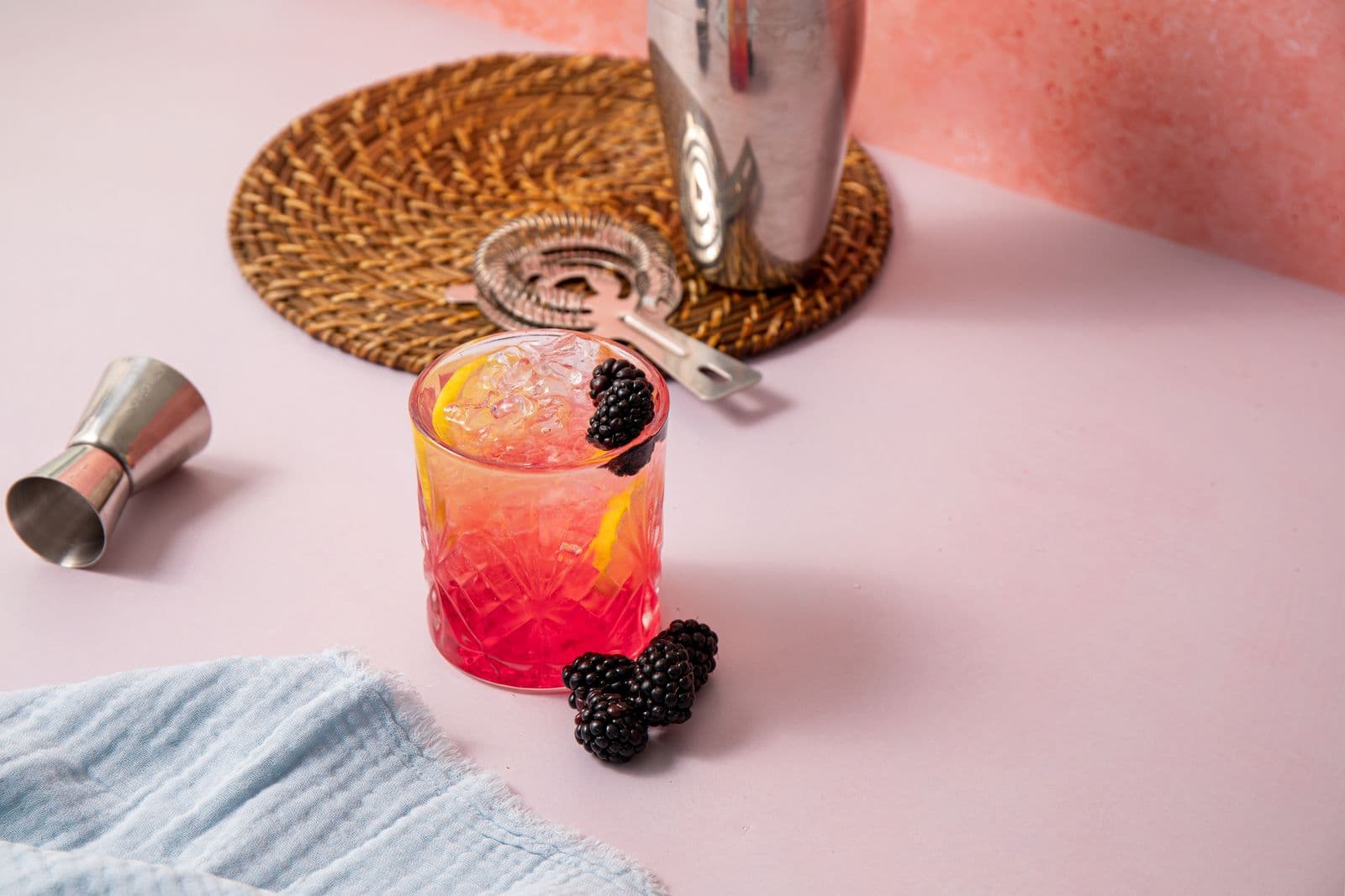 A stainless steel shaking tin, a Hawthorne strainer and a double jigger beside a blackberry cocktail on a pale pink counter