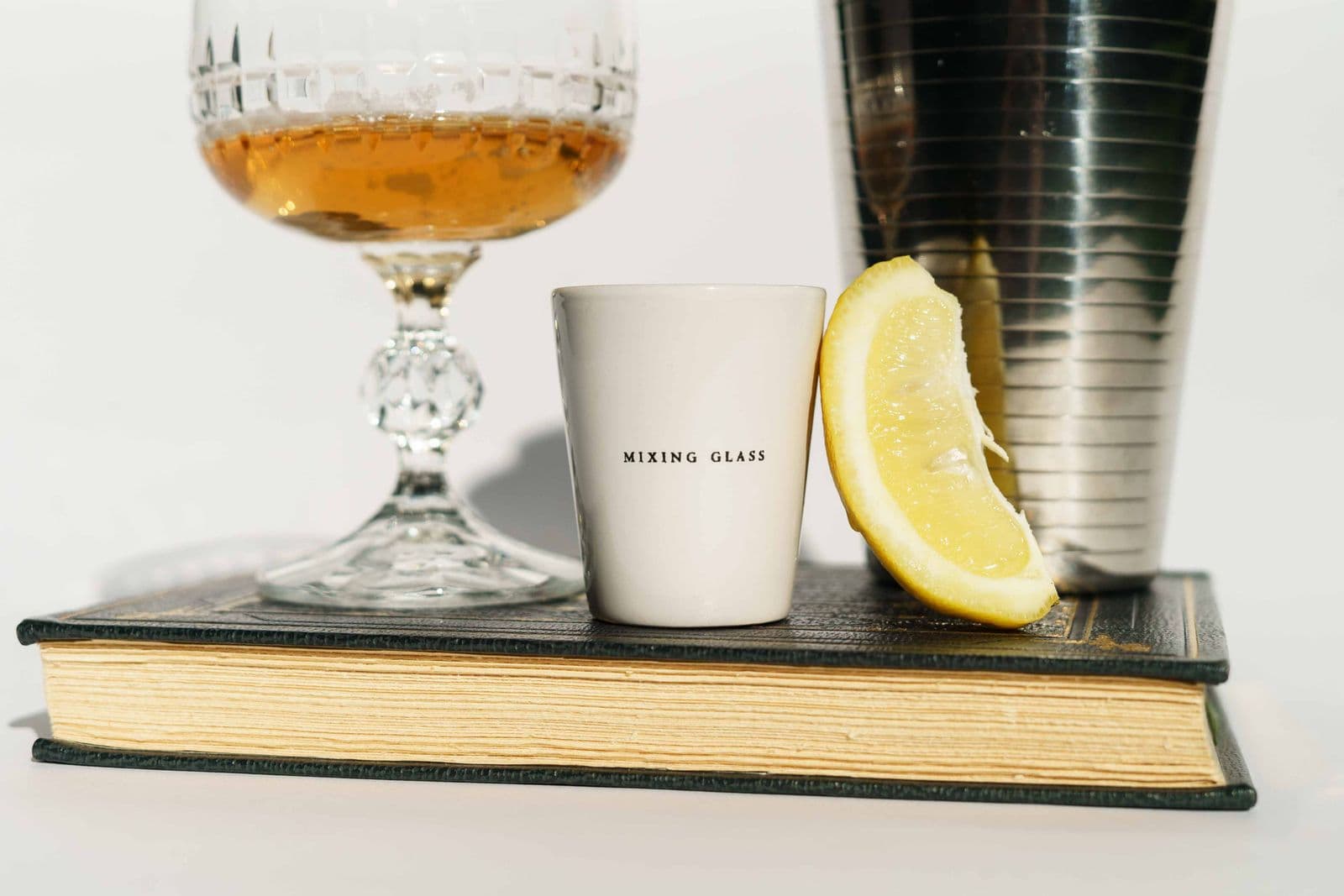 A hardback book topped with a shaker tin, a jigger, a lemon wedge and a glass of amber spirit
