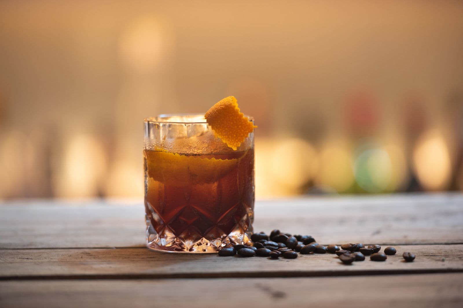 A dark coffee cocktail with an orange twist beside scattered coffee beans