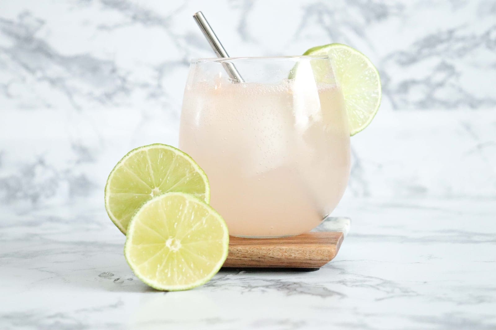 A pale citrus drink with lime slices and a metal straw on a marble surface