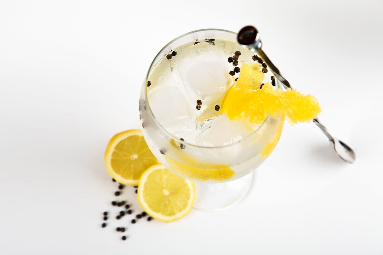 A gin and tonic in a stemmed balloon glass with lemon slices and peppercorns on white