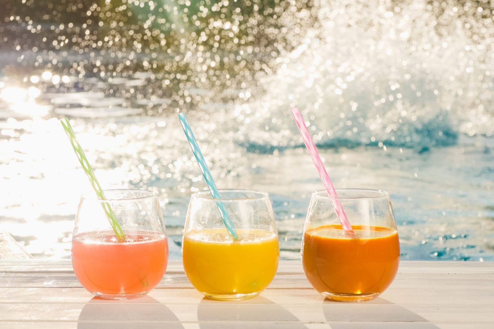 Three stemless tumblers of fruit juice with striped straws beside a sunlit pool